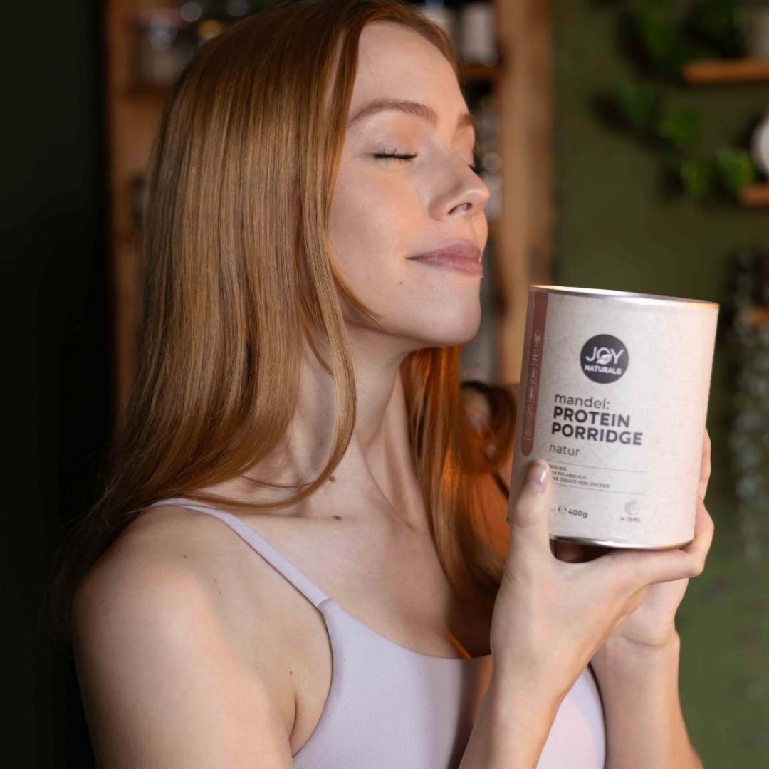 Frau die am herrlich duftenden Mandel Protein Porridge von JOY NATURALS riecht.
